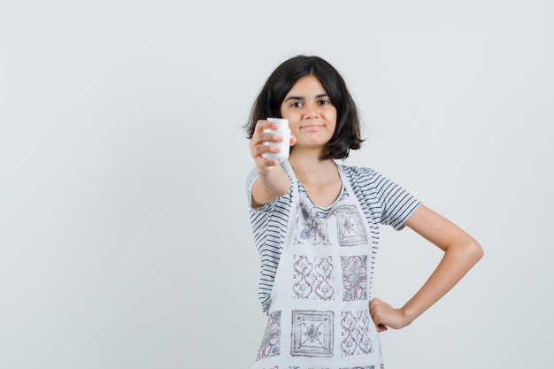 Niña que presenta una botella de píldoras en camiseta, delantal y parece confiada.