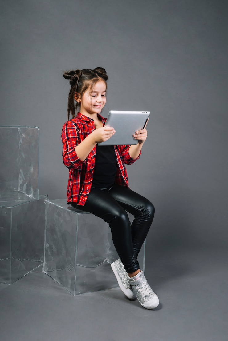 Foto gratuito niña que se sienta en el bloque que mira la tableta digital contra fondo gris