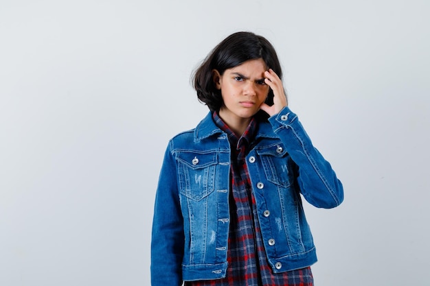 Niña rascándose la cabeza, pensando en algo en camisa a cuadros y chaqueta de mezclilla y mirando pensativo, vista frontal.