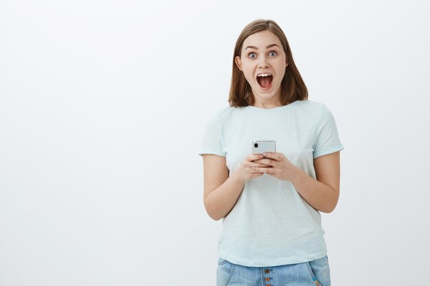 La niña recibió una invitación a una fiesta increíble a través de mensajes en Internet con un teléfono inteligente mirando sorprendida y encantada con la boca abierta de la emoción mirando felizmente sobre la pared gris