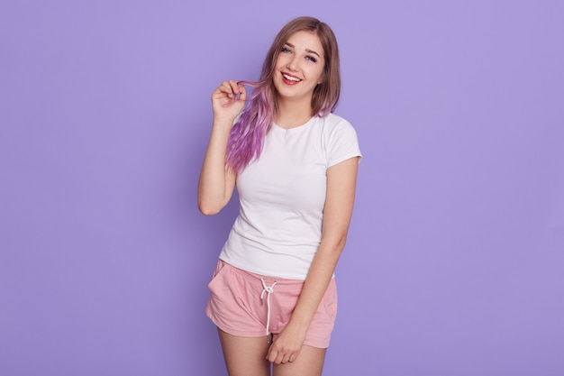 Niña riendo feliz en camiseta blanca y rosa corta con expresión coqueta, manteniendo los dedos en su cabello lila, posando aislada sobre pared púrpura.