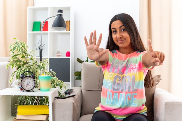 Niña en ropa casual feliz y segura mostrando el número seis sonriendo sentado en la silla en la sala de luz