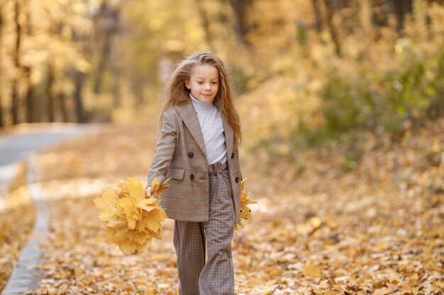 Niña con ropa de moda caminando en el parque de otoño. Chica sosteniendo hojas amarillas. Vestida de traje marrón con chaqueta.