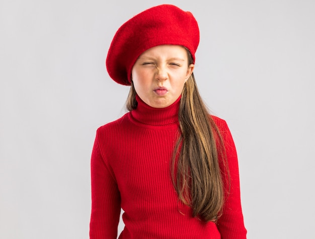 Niña rubia con el ceño fruncido vistiendo boina roja mirando al frente aislado en la pared blanca con espacio de copia