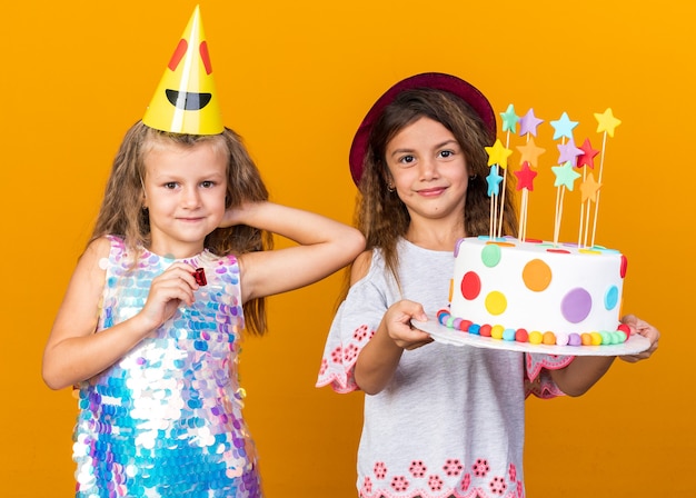 Niña rubia complacida con gorro de fiesta sosteniendo un silbato y de pie con una niña caucásica con sombrero morado y sosteniendo un pastel de cumpleaños aislado en una pared naranja con espacio de copia