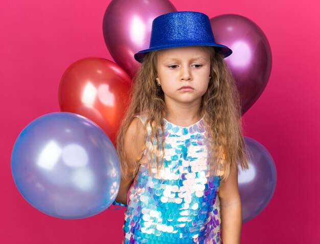 Niña rubia decepcionada con gorro de fiesta azul de pie con globos de helio aislado en la pared rosa con espacio de copia