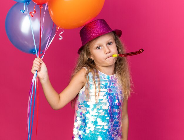 Niña rubia decepcionada con gorro de fiesta púrpura sosteniendo globos de helio y soplando silbato de fiesta aislado en la pared rosa con espacio de copia