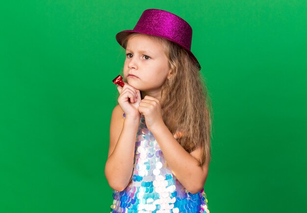 Niña rubia despistada con gorro de fiesta púrpura sosteniendo un silbato de fiesta y mirando al lado aislado en la pared verde con espacio de copia