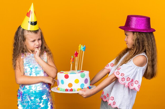 Niña rubia disgustada con gorra de fiesta mirando a la niña caucásica con sombrero de fiesta púrpura sosteniendo pastel de cumpleaños aislado en la pared naranja con espacio de copia