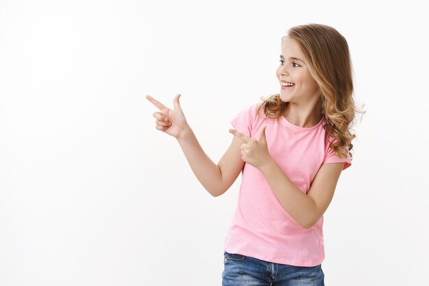 La niña rubia feliz sonriente alegre despreocupada gire a la izquierda que señala el espacio de la copia de las pistolas del dedo, saludando a los amigos de la escuela, sonriendo encantado. Niño echa un vistazo a la genial promoción de copyspace, pared blanca