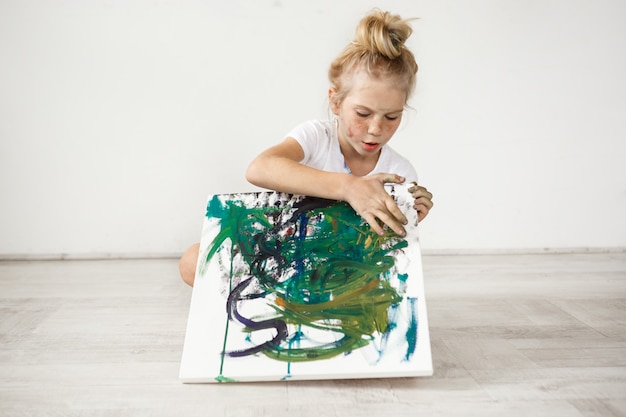 Niña rubia con hairbn y pecas con camiseta blanca ocupada con su foto. Linda y adorable niña sentada en un piso con lienzo de colores sobre sus rodillas.