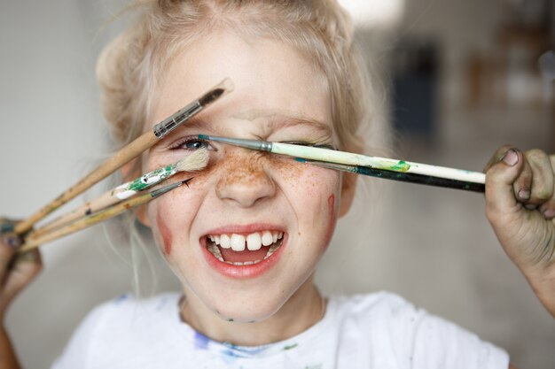 Niña rubia de humor juguetón con pintura en su cara pecosa y ojos azules que cubren su rostro con pinceles