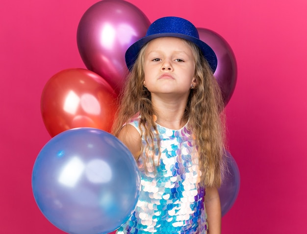 Niña rubia molesta con gorro de fiesta azul de pie con globos de helio aislado en la pared rosa con espacio de copia