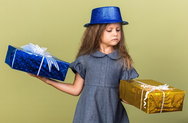 Niña rubia molesta con gorro de fiesta azul sosteniendo y mirando cajas de regalo aisladas en la pared verde oliva con espacio de copia