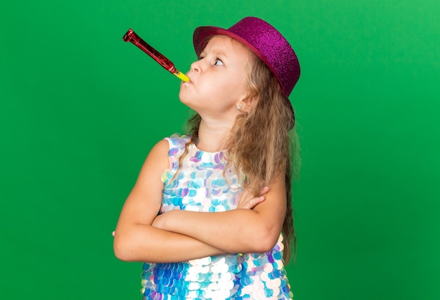 Niña rubia molesta con gorro de fiesta púrpura de pie con los brazos cruzados soplando el silbato de fiesta y mirando al lado aislado en la pared verde con espacio de copia