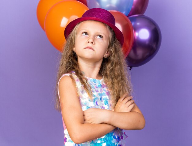Niña rubia molesta con sombrero de fiesta violeta de pie con los brazos cruzados frente a globos de helio mirando hacia arriba aislado en la pared púrpura con espacio de copia