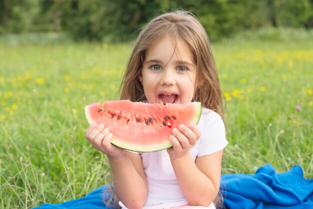 Niña rubia que come la rebanada de la sandía en el parque