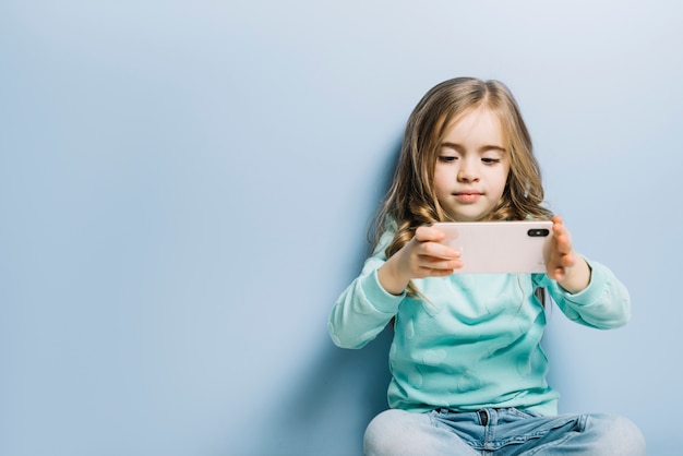 Niña rubia que se sienta contra fondo azul que mira el vídeo en el teléfono móvil