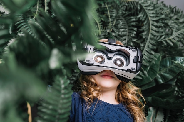 Niña rubia sonriente que usa las auriculares virtuales que se colocan entre la planta verde