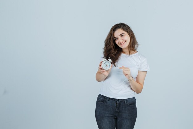 Niña señalando el despertador en camiseta, jeans y mirando alegre. vista frontal.