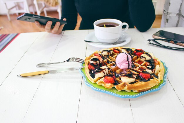 Niña sentada caffe comiendo desayuno waffle con salsa de chocolate, rodajas de plátano y fresas en un plato de cerámica verde y fotografió su desayuno