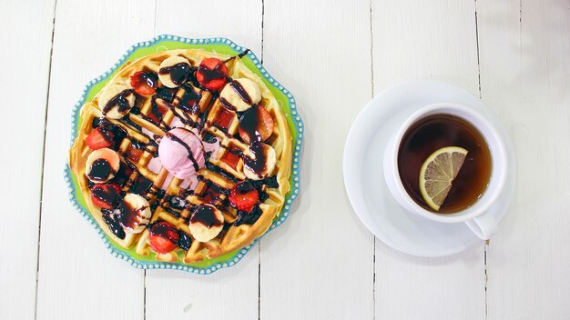 Niña sentada caffe comiendo desayuno waffle con salsa de chocolate, rodajas de plátano y fresas en un plato de cerámica verde y fotografió su desayuno