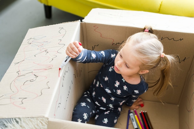 Foto gratuita niña sentada en una caja de cartón y decorarla con un rotulador