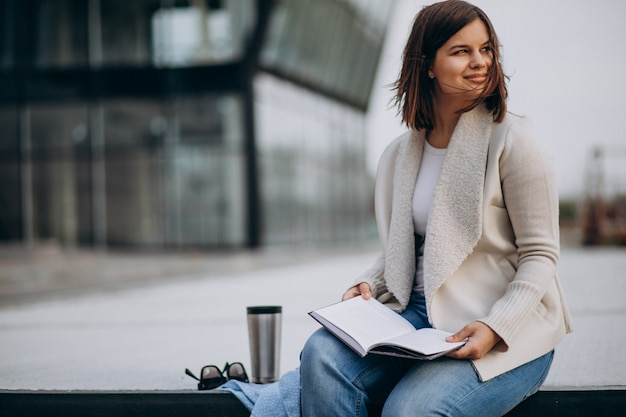 Foto gratuita niña sentada leyendo un libro y tomando café fuera de la calle