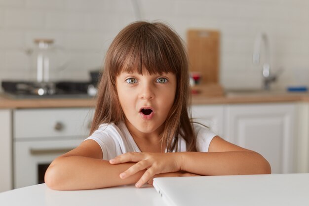 Niña sentada a la mesa con la tapa del regazo cerrado en casa. Linda chica mirando a cámara con la boca abierta, sorprendida, niña de cabello oscuro asombrada después de ver contenido sorprendido.