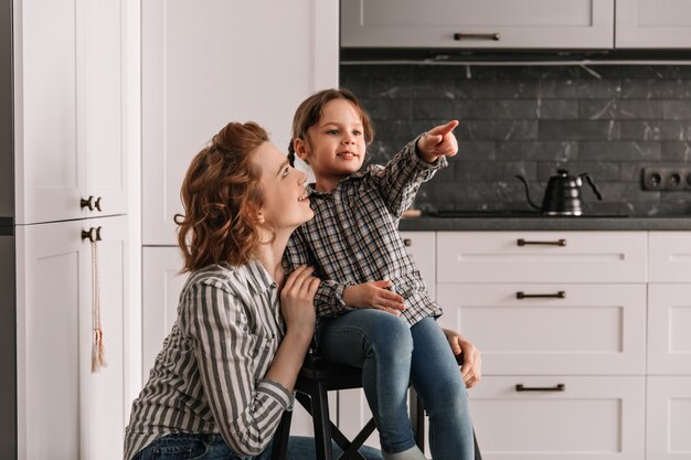 La niña se sienta en una silla y señala con el dedo a un lado. Mamá e hija posando en la cocina.