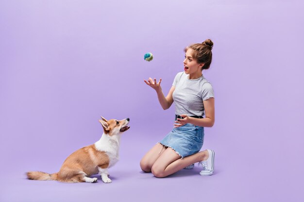 La niña se sienta en el suelo y juega con el perro. Mujer joven feliz en zapatillas blancas posando con pelota y corgi sobre fondo púrpura.