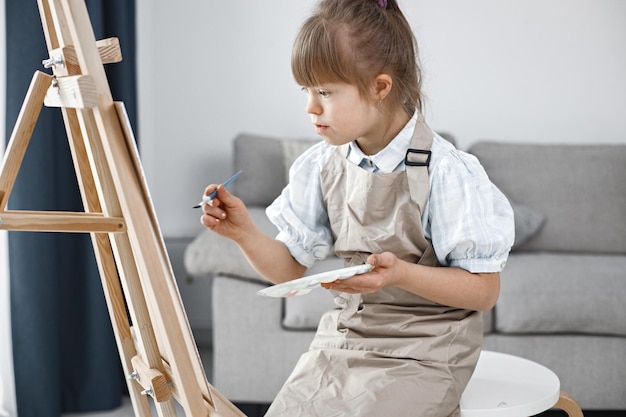 Foto gratuita niña con síndrome de down con delantal beige pintando en un caballete