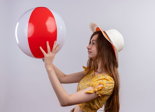 Foto gratuita niña con sombrero levantando la pelota de playa y mirándola de pie en la vista de perfil en la pared blanca aislada