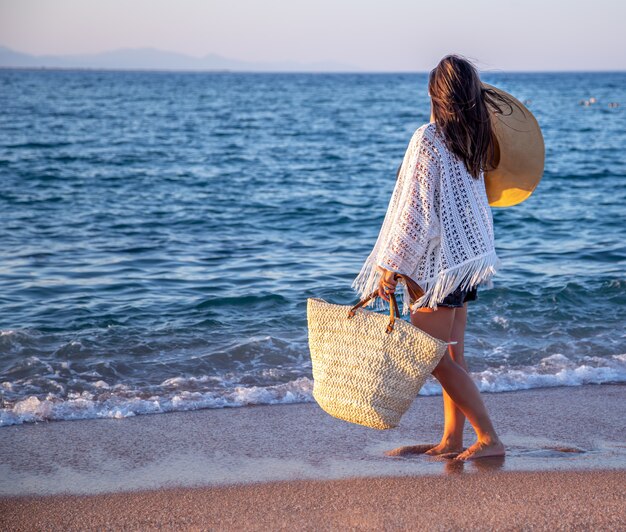 Una niña con un sombrero en las manos y una bolsa de mimbre camina por la orilla del mar. Concepto de vacaciones de verano.