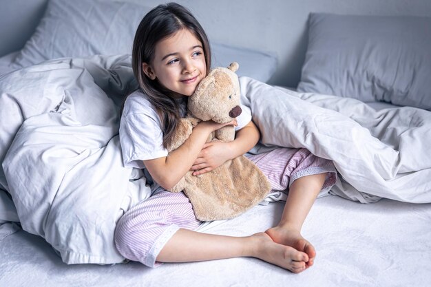 Niña soñolienta con su osito de peluche favorito en la cama por la mañana