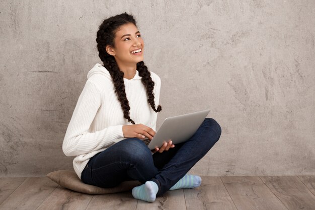 Niña sonriendo, sosteniendo portátil, sentado en el piso sobre pared beige