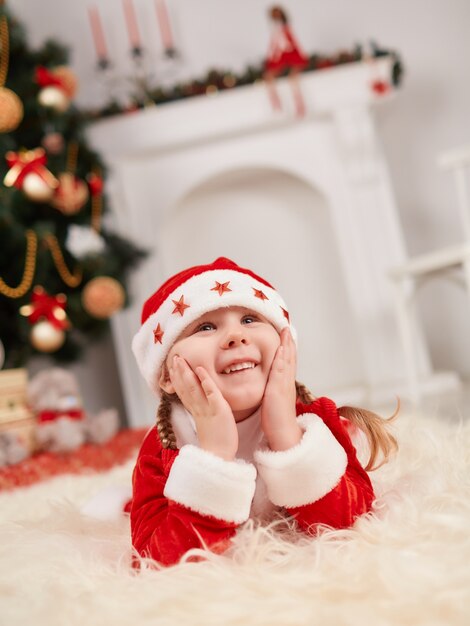 Niña sonriendo vestida de papa noel tumbada en el suelo con las manos en la cara