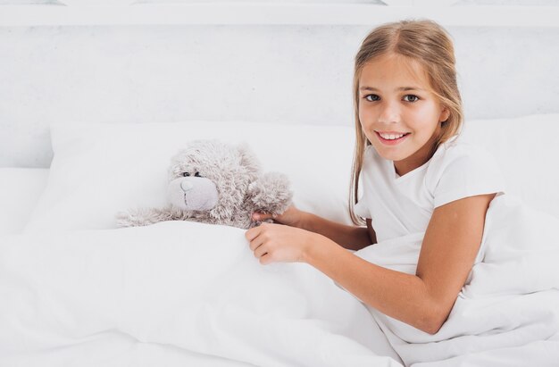 Niña sonriente de alto ángulo quedarse en la cama con su osito de peluche