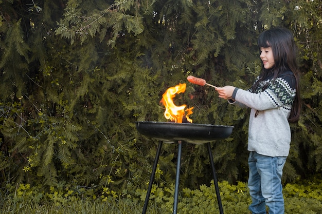 Niña sonriente asar salchichas en barbacoa portátil al aire libre