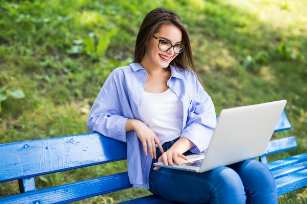 Foto gratuita niña sonriente en camiseta azul sentarse en el banco en el parque y usar su nueva computadora portátil