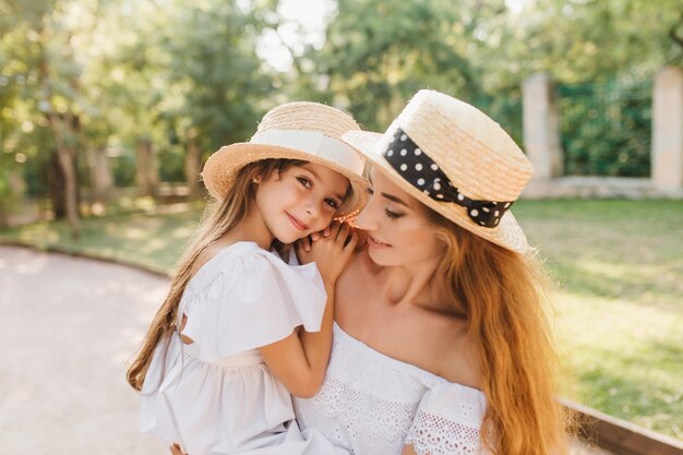 Niña sonriente cansada en canotier de paja descansando sobre las manos de la madre después de una larga caminata en el parque. Retrato al aire libre de la hija hermosa alegre que lleva de la mujer en vestido blanco que abraza suavemente su cuello.