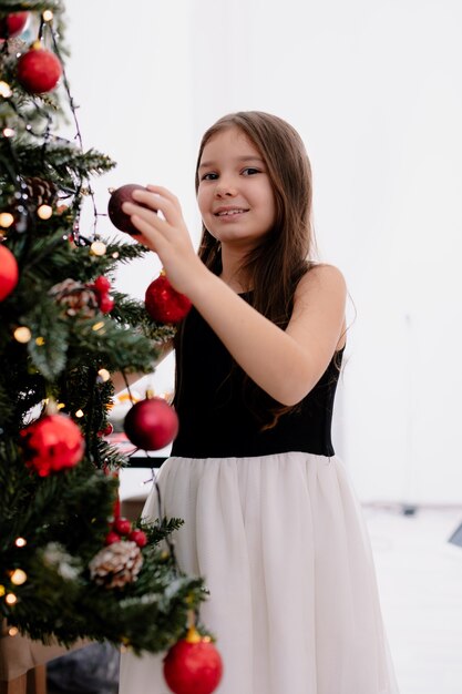 Niña sonriente en casa en la época navideña decorar el árbol de navidad en el salón sosteniendo adornos navideños