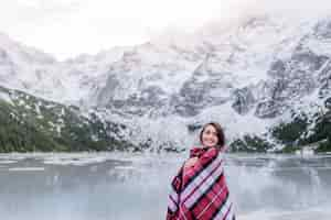 Foto gratuita niña sonriente cubierta con una manta cálida está de pie cerca del lago en las montañas, joven viajero