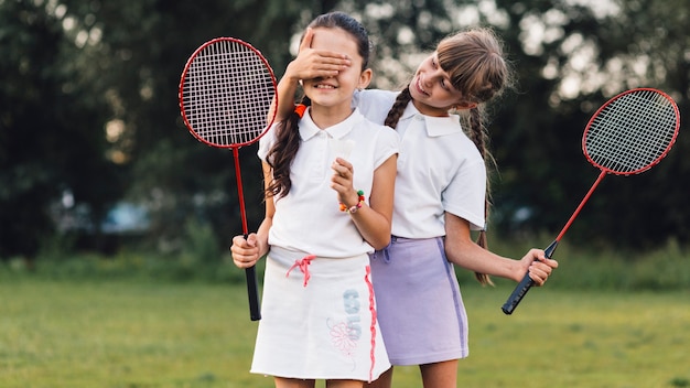 Niña sonriente cubriendo sus ojos amiga sosteniendo bádminton