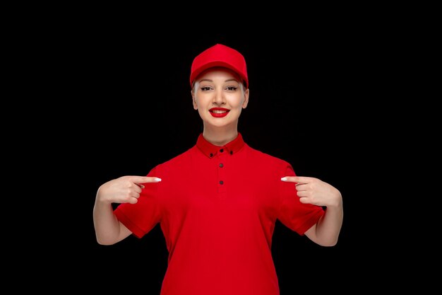 Niña sonriente del día de la camisa roja apuntándose a sí misma con una gorra roja con camisa con lápiz labial