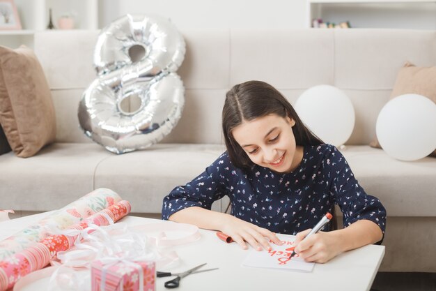 Niña sonriente en el día de la mujer feliz escribe en la tarjeta de felicitación sentado en el piso detrás de la mesa de café con regalos en la sala de estar