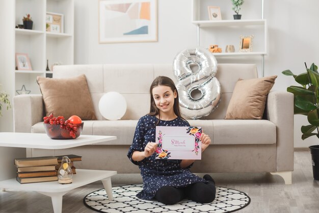 Niña sonriente en el día de la mujer feliz sentada en el piso sosteniendo la tarjeta de felicitación en la sala de estar