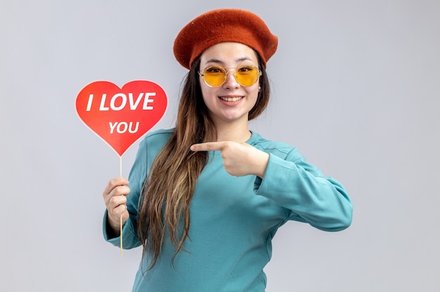 Niña sonriente en el día de San Valentín con sombrero con gafas sosteniendo y puntos en el corazón rojo en un palo con te amo texto aislado sobre fondo blanco.