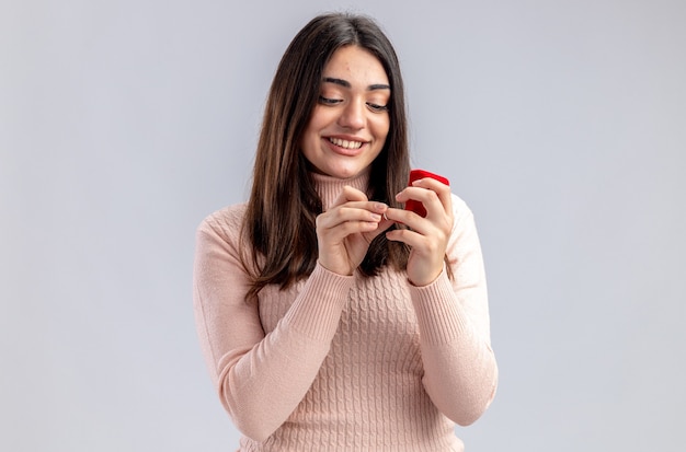 Niña sonriente en el día de San Valentín sosteniendo y mirando el anillo de bodas aislado sobre fondo blanco.