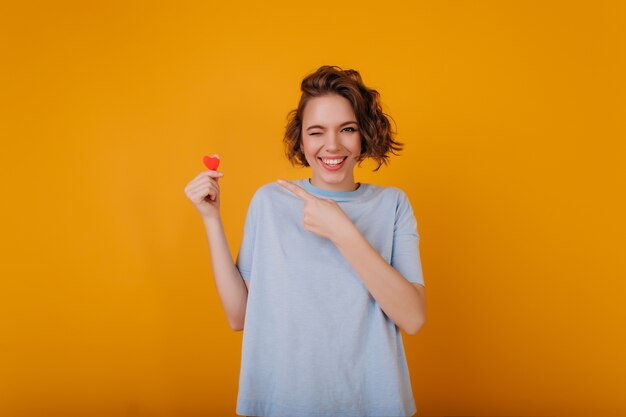 Niña sonriente de ensueño con el corazón en la mano divirtiéndose durante la sesión de fotos. Maravillosa mujer rizada escalofriante en estudio en el día de San Valentín.